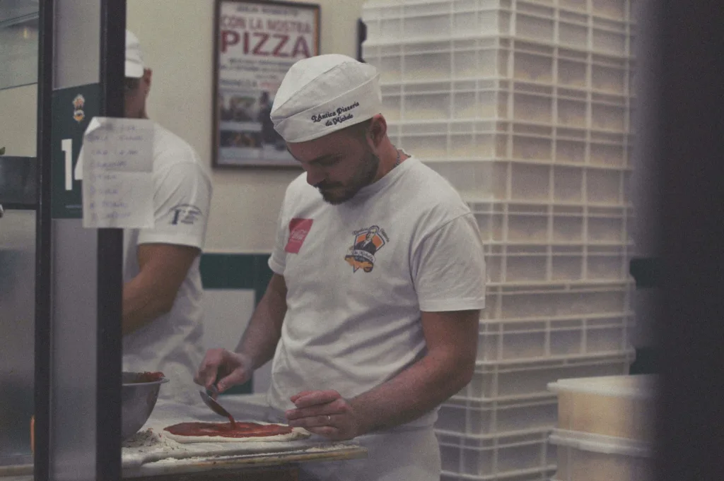 Pizza chef spreading sauce on dough in Napoli restaurant kitchen.
