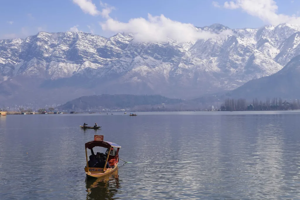 Peaceful view of Dal Lake in Srinagar surrounded by snowcapped Himalayas, featuring traditional boats.
